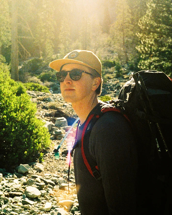 Man wearing harvest gold flat-brim Adventure Hat hiking on a sunlit rocky trail with a backpack