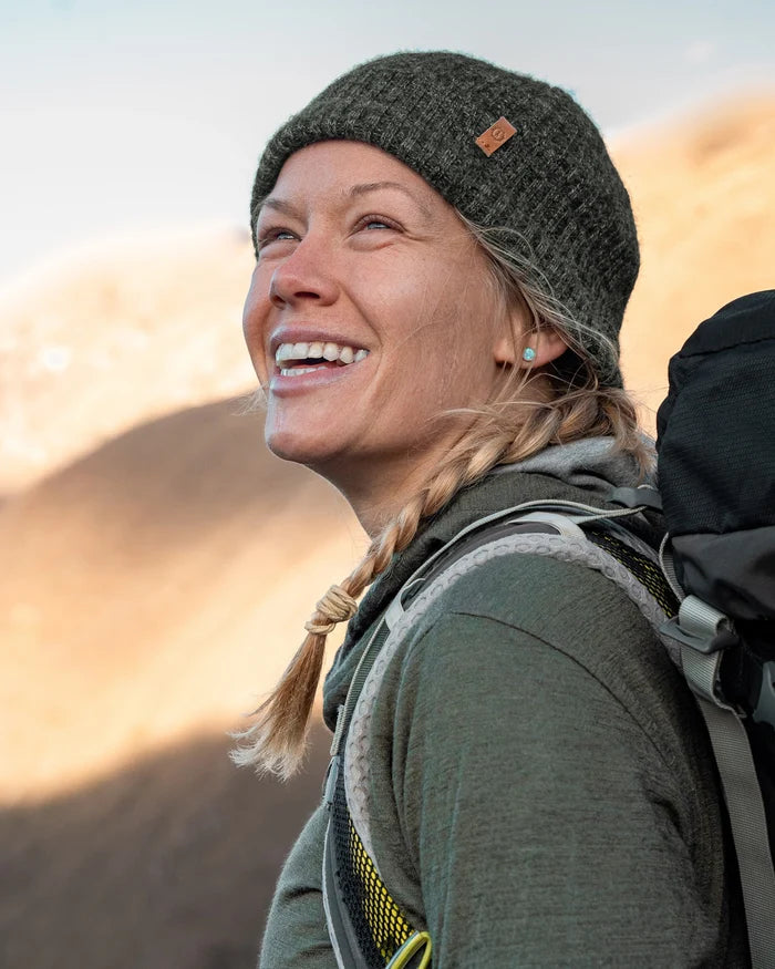 Smiling woman hiker wearing a dark green marled beanie and backpack in a sunlit mountain landscape