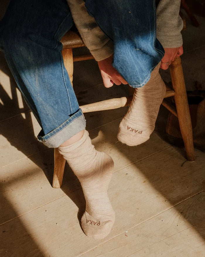 One pair of feet wearing Timber socks with PAKA branding, sitting on a wooden stool.
