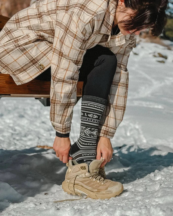 Model pulling on charcoal Inca-pattern crew socks while putting on tan hiking boot in snowy outdoor setting
