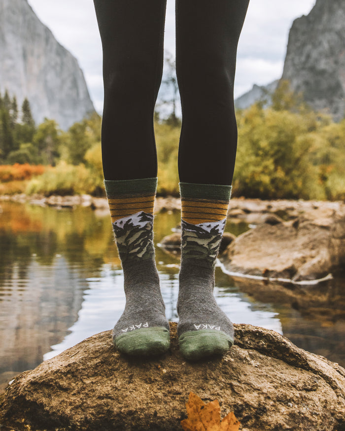 Front view of Essential Mountain crew socks showcasing mountain graphic and cushioned toe on a rock by a river and peaks