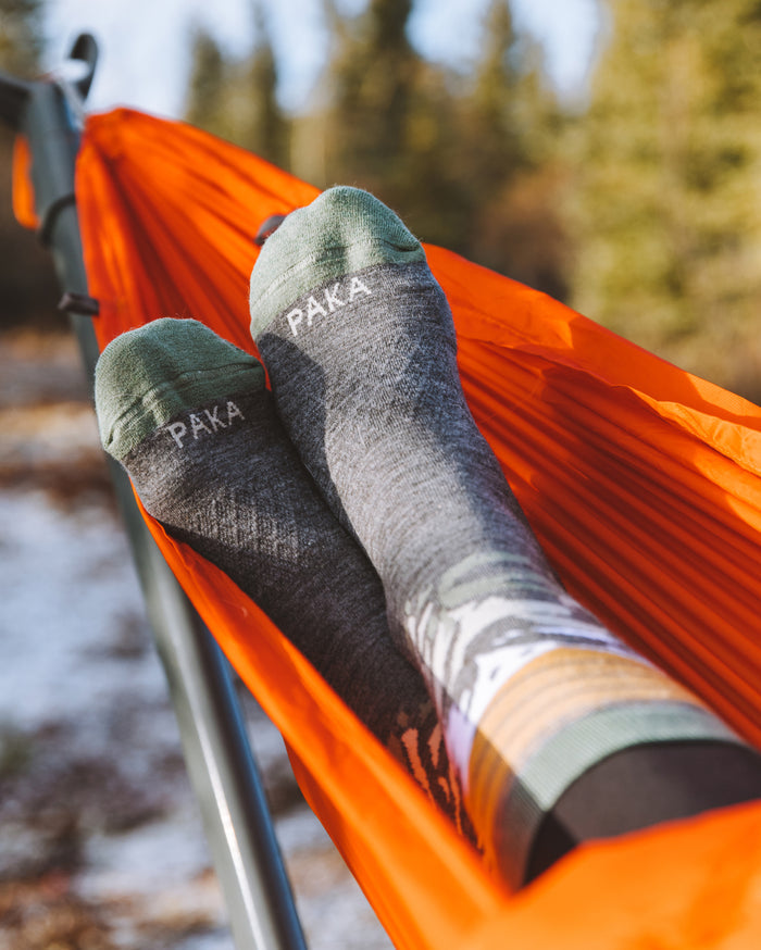 Person’s feet wearing gray-and-green mountain crew socks with ‘PAKA’ on the toes, resting in an orange hammock outdoors