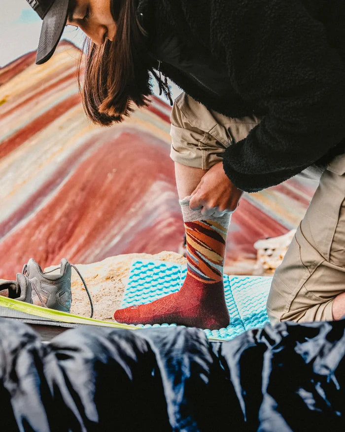 Hiker pulling on a Rainbow Mountain crew sock while kneeling on a blue foam mat with colorful striped mountain in the background