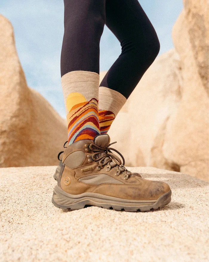 Person standing on a rock wearing Rainbow Mountain crew socks with multicolored mountain pattern and hiking boot in a sunlit desert setting