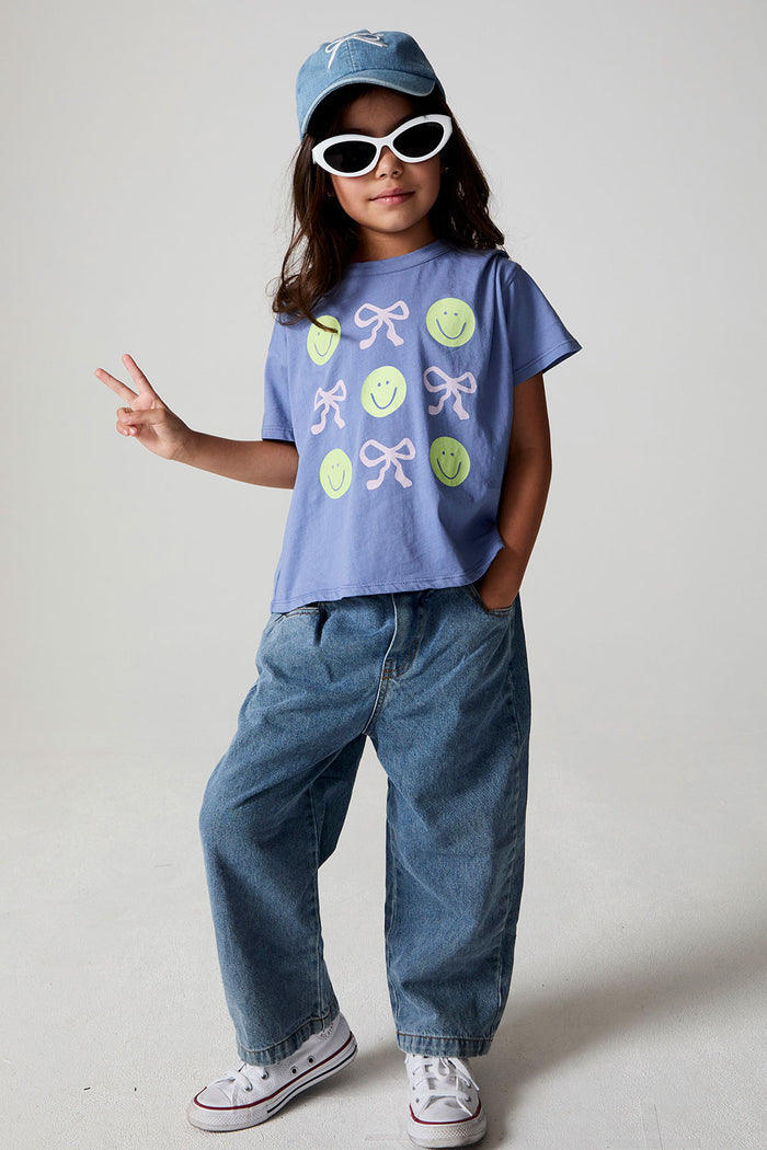 Full body front view of a girl wearing the Chaser Happy Bows Tee with sunglasses and a hat on a neutral background
