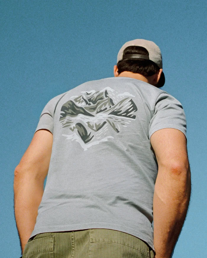 Person wearing a gray PAKA "Love Nature Back" t-shirt with a graphic design on the back against a clear blue sky.