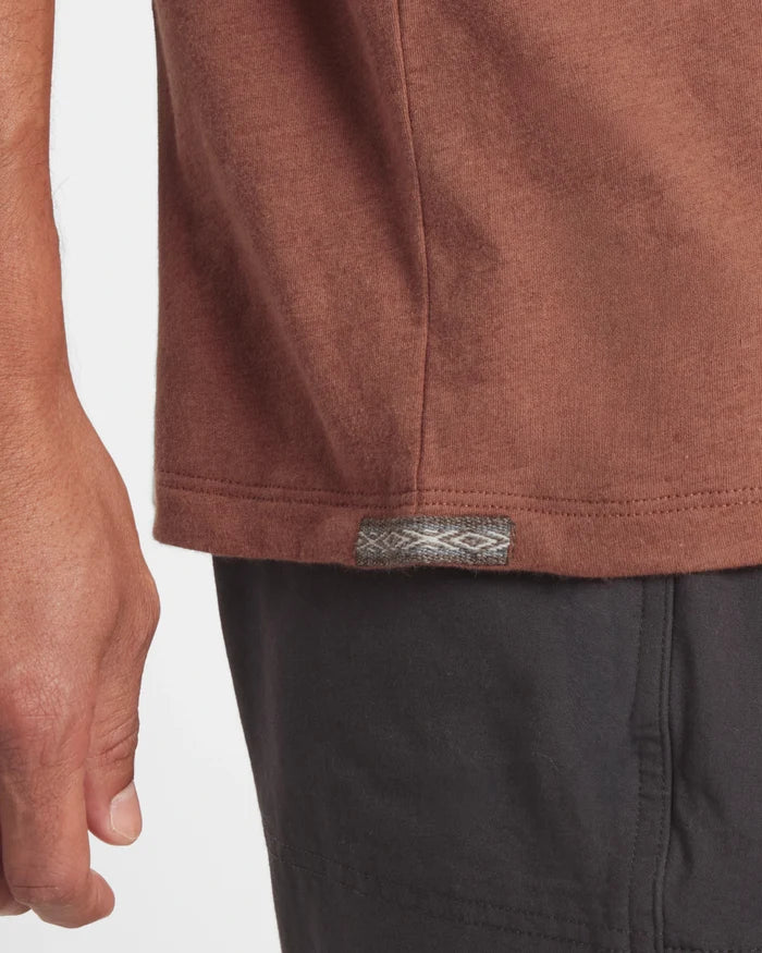 Close up of Men’s Essential Tee hem in cinnamon showing side seam and woven logo tab with model’s hand and black shorts