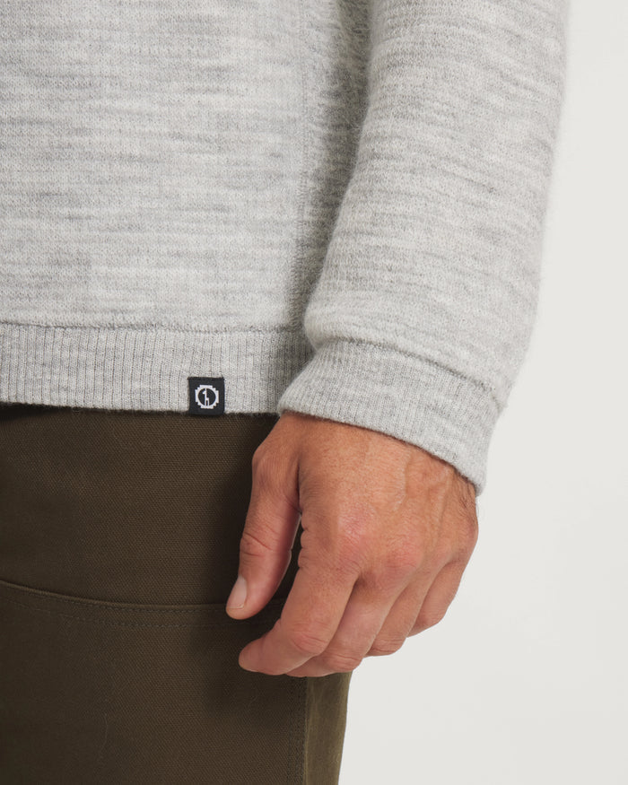 Close-up of Men’s Mountain Crew sweater showing ribbed hem, rolled cuff, heather light grey knit and small black logo tag on white background