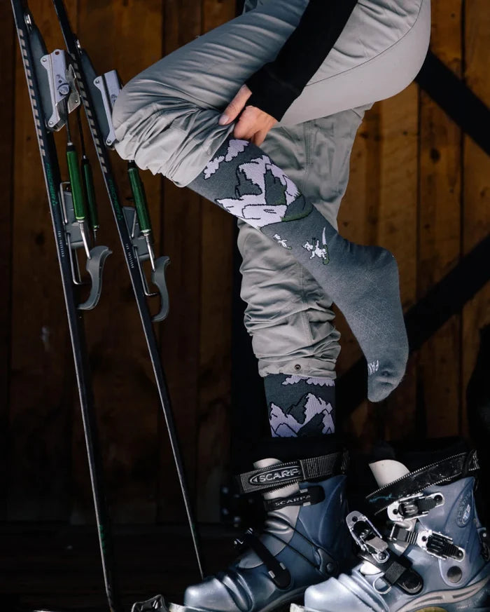 Side view of Snow Sebastian indigo ski socks on a model showing leg compression and cushioned foot with ski boots and wooden cabin background