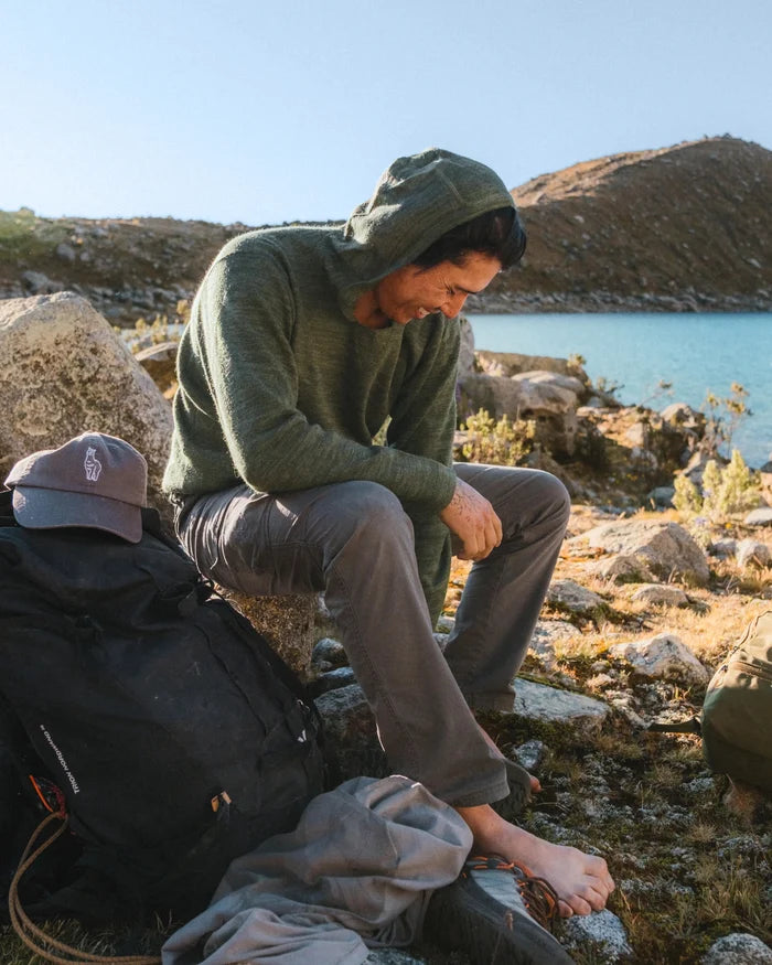 Man wearing eucalyptus alpaca hoodie sitting on rocks by a mountain lake during a relaxed outdoor hike