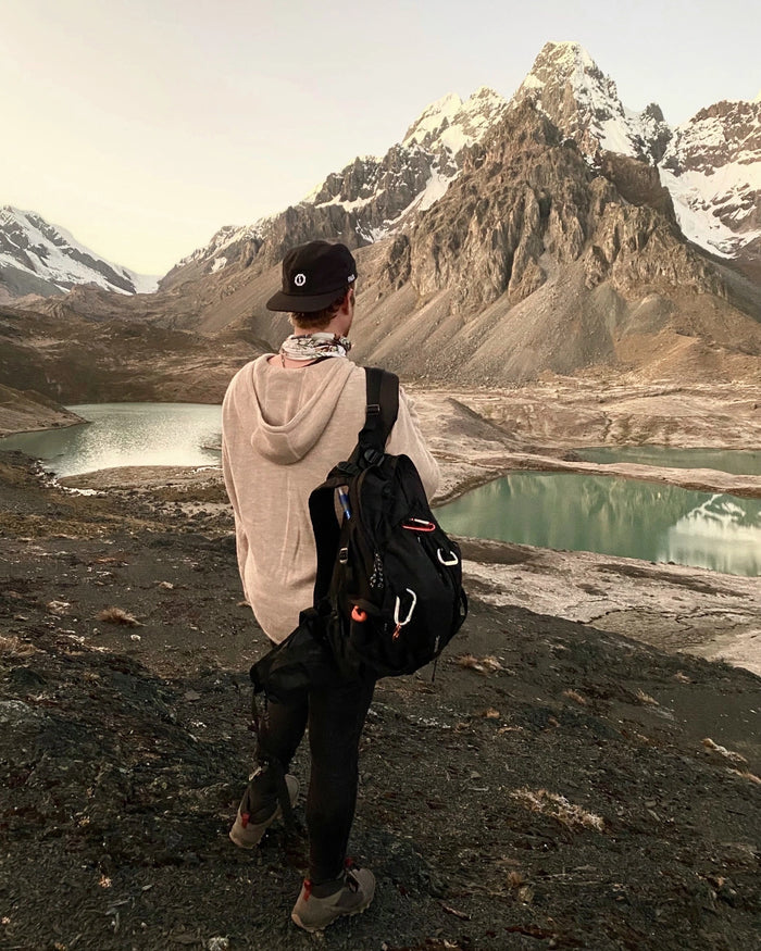 Back view of The Hoodie in oat showing relaxed fit with hood down and backpack at mountain lake