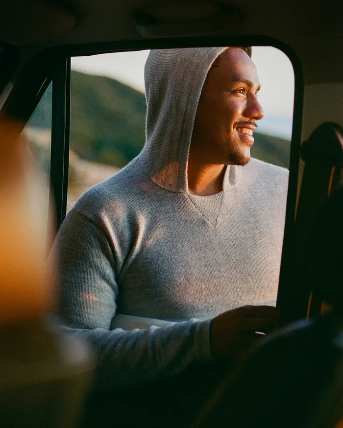 Man smiling and looking out a car window wearing a light grey alpaca hoodie, relaxed fit, coastal sunset background