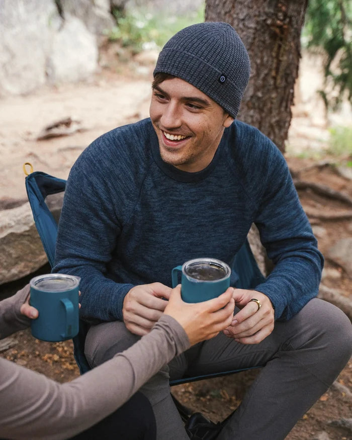 Man wearing a navy alpaca crewneck sweater in regular fit smiling and sharing a mug while seated at a campsite