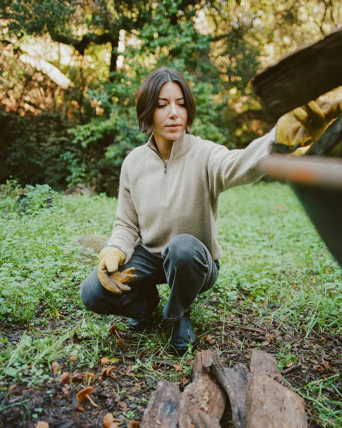 Woman crouching outdoors wearing a pebble alpaca quarter-zip sweater in a regular fit, reaching toward a wheelbarrow in a wooded yard