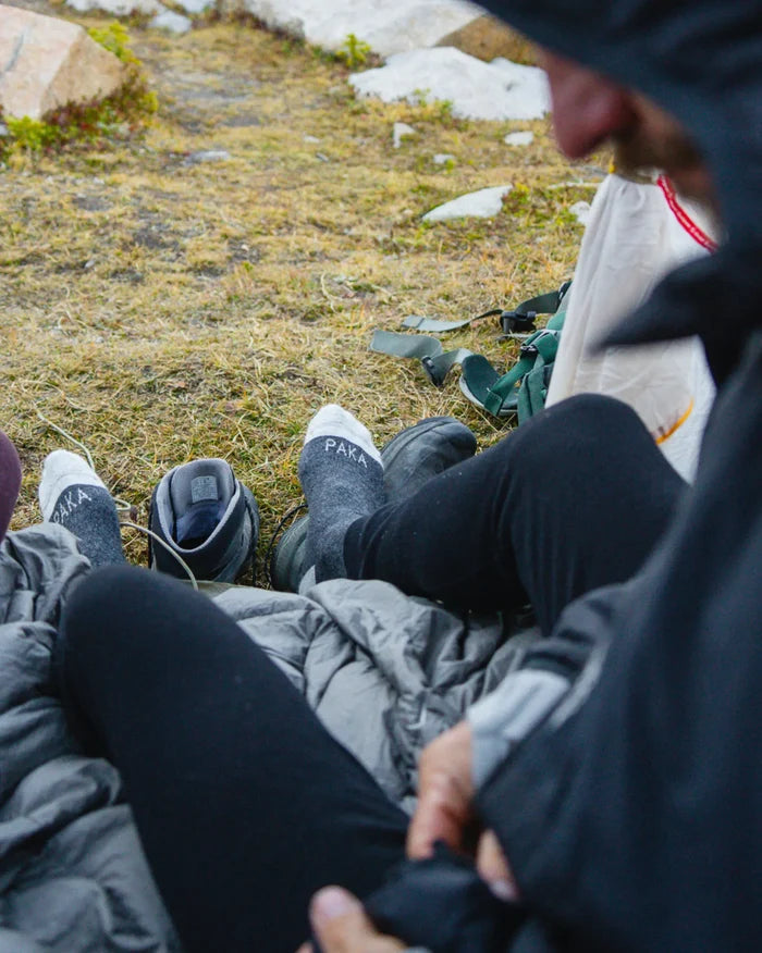 Two people sitting on a sleeping bag at a campsite, wearing grey Thermal Condór crew socks with PAKA logo on the toes