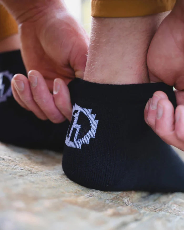 Close-up of black Trail 1/4 crew sock on heel showcasing white PAKA logo and elastic cuff on rocky ground
