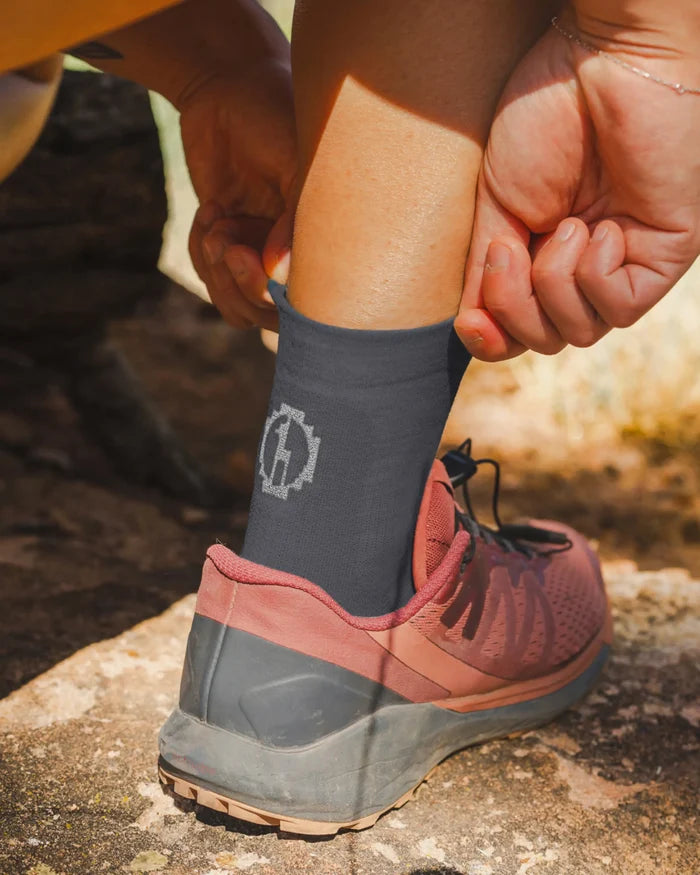 Model adjusting charcoal-blue Trail 1/4 crew sock on rocky trail, showing ankle fit