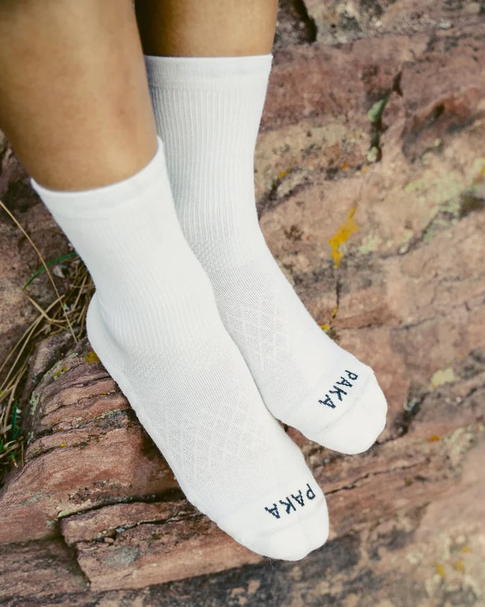 Angled top view of white Trail 3/4 crew socks worn on a model, ribbed cuff and textured footbed, PAKA logo on toes, on rock