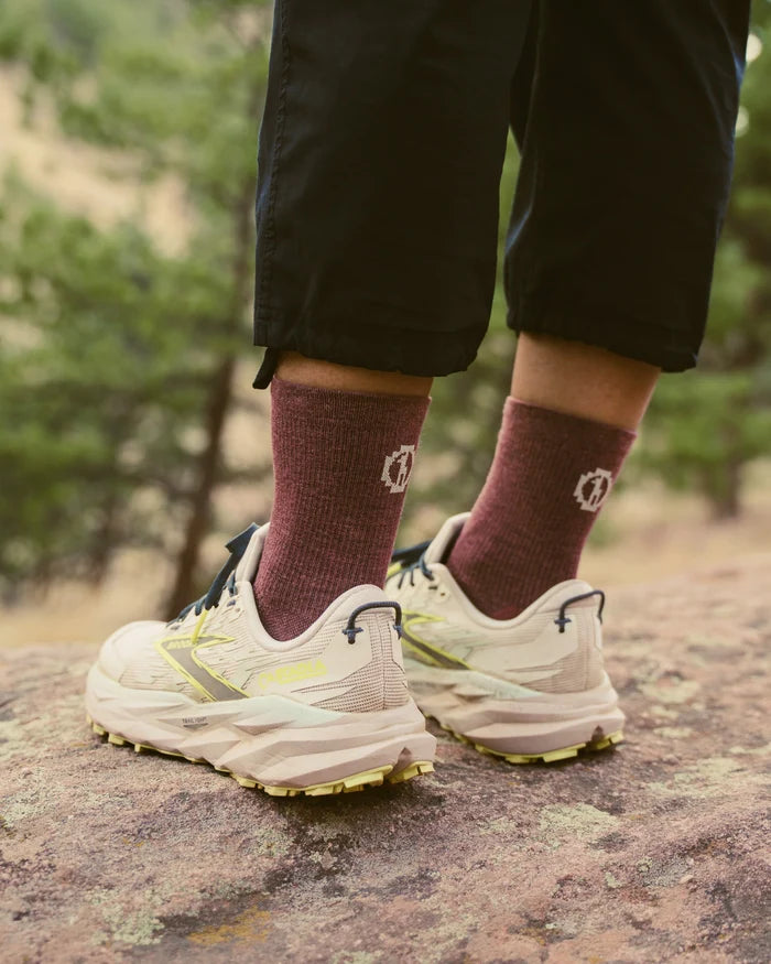 Model standing on a rock wearing maroon 3/4 crew trail socks and beige trail running shoes in a wooded outdoor setting