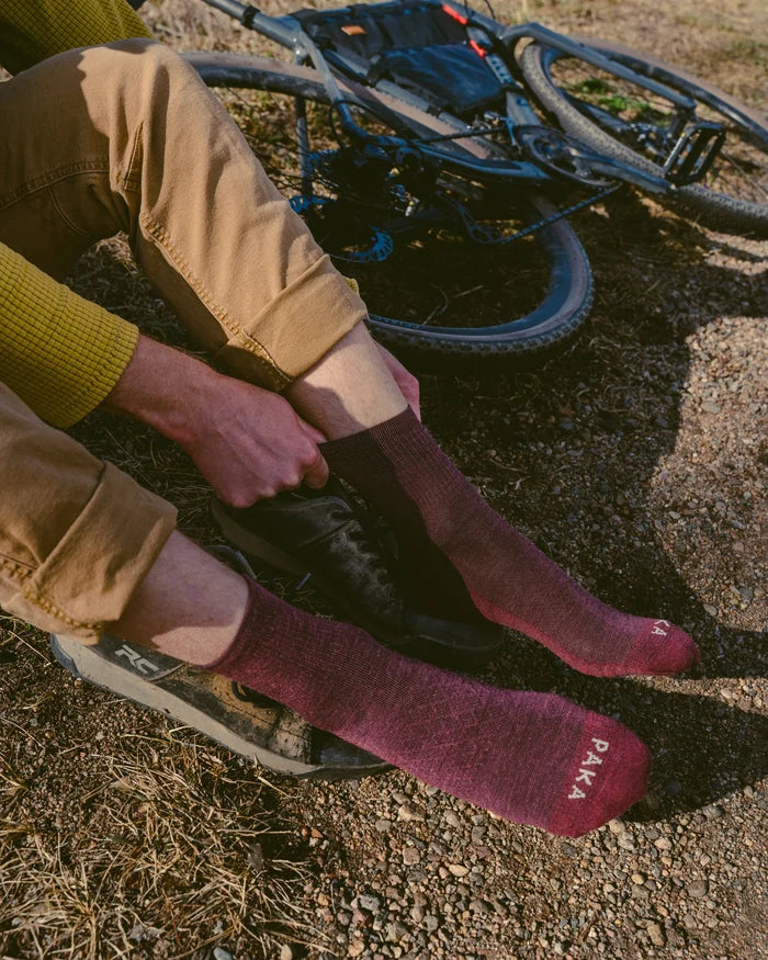 Model pulling on maroon 3/4 crew PAKA hiking socks showing PAKA logo on toes, sitting by a gravel trail with a mountain bike
