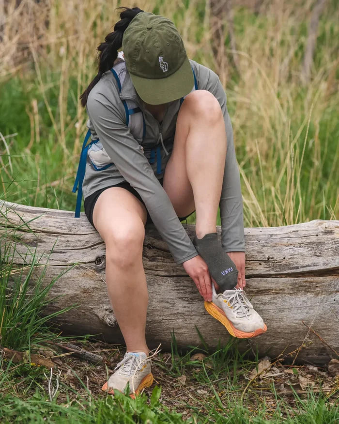 Woman sitting on a log putting on charcoal-blue Trail Ankle Socks made from alpaca, wearing shorts and hiking shoes on a grassy trail