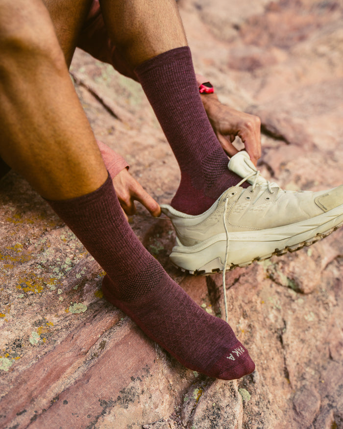 Angled view of Trail Crew Socks in burgundy showing crew height, textured knit and reinforced toe with PAKA logo on rocky background