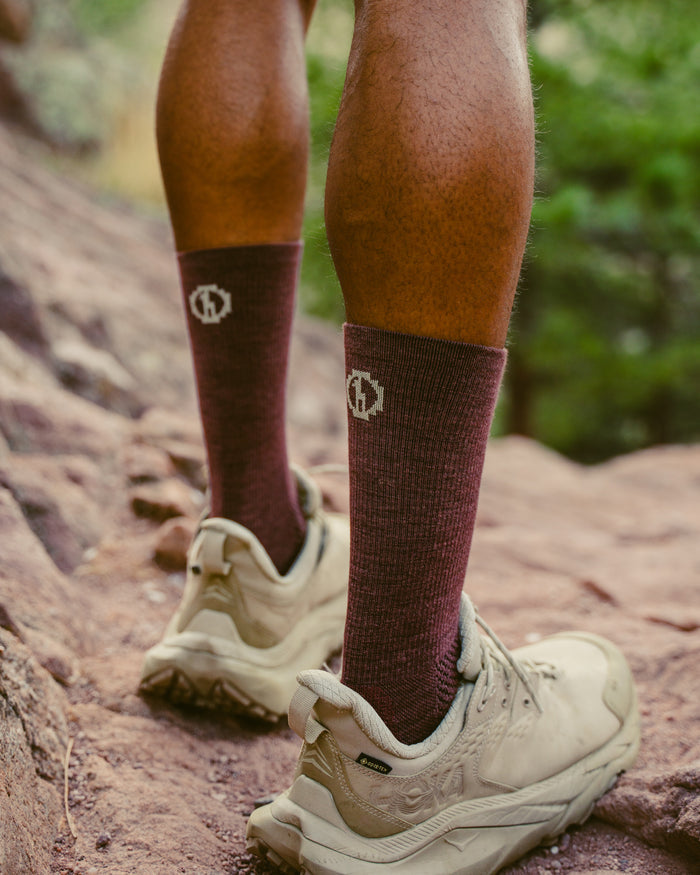 Rear view of burgundy Trail Crew socks on a hiker showing ribbed crew height and white embroidered logo on a rocky trail