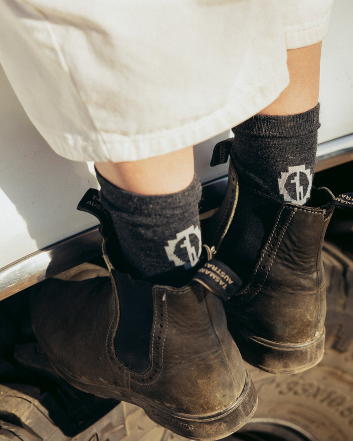 Rear close-up of Trail Crew Socks in black on a model’s ankles, showing crew length, ribbed cuff, PAKA logo, worn with leather boots