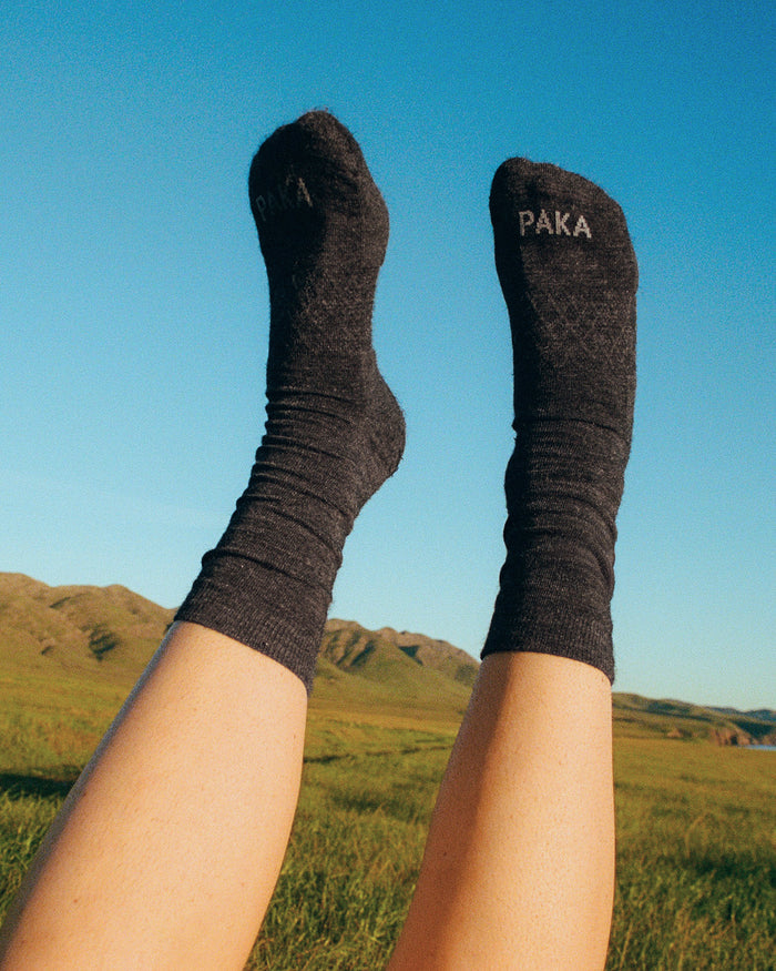 Legs wearing charcoal mid-calf Trail Crew alpaca socks raised over grassy hills under blue sky