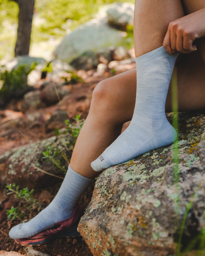 Side view of light blue Trail Crew socks on a seated model, showing calf-height ribbed knit and cushioned sole on a rocky trail